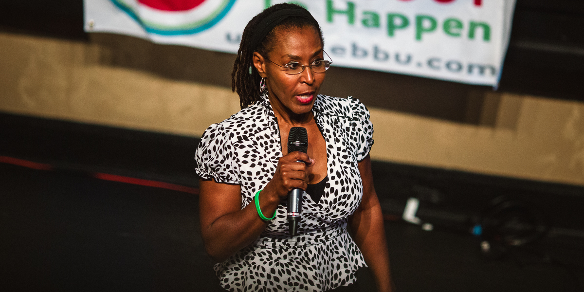 A woman in a black and white patterned dress speaks into a microphone while standing on stage at the cuban club ybor city. A blurry banner is visible in the background.