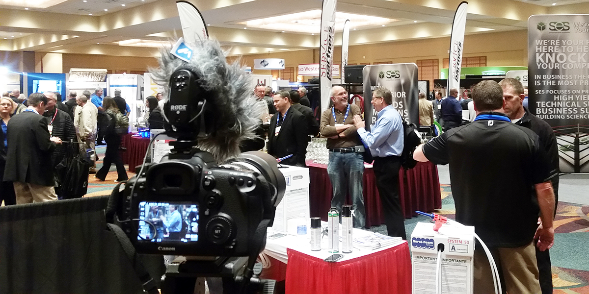 A camera on a tripod in focus at a busy exhibition hall, capturing a scene of various booths and attendees engaged in conversations.