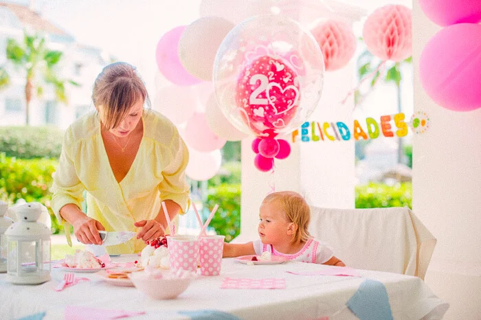 A woman in a yellow shirt is cutting cake at a table while a toddler looks on. The table is decorated with pink party items, and a large balloon with "2" and "felicidades" is in the background.