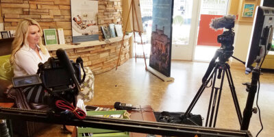 Woman speaking in an indoor studio with cameras, tripods, microphones, and promotional banners.