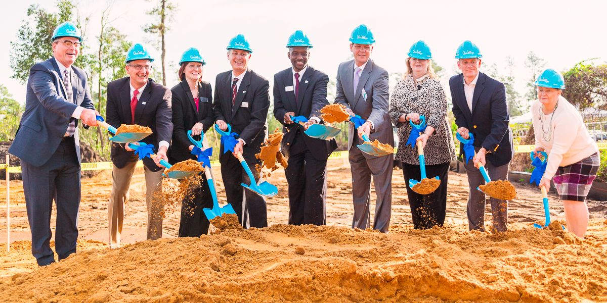 A group of nine people in business attire and blue helmets participate in a groundbreaking ceremony, holding shovels as they dig into a pile of dirt.