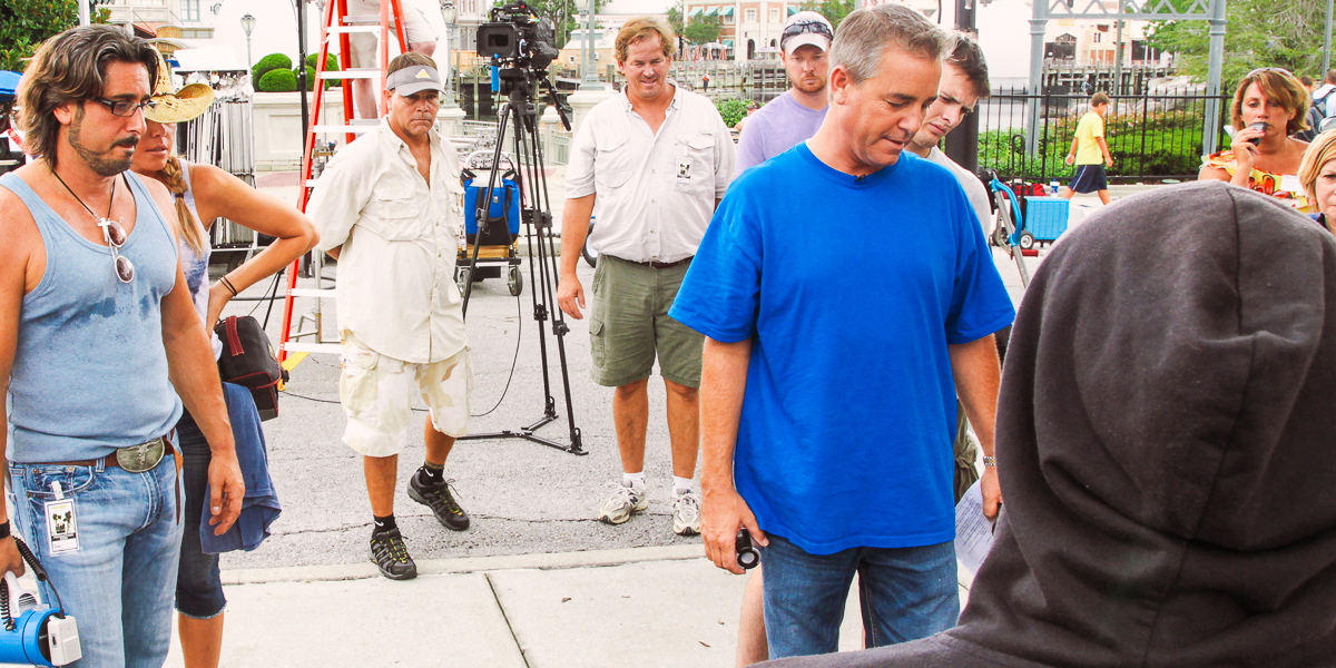 A group of people, including some in casual clothing and one in a hooded sweatshirt, are gathered outdoors near film equipment at universal studios orlando. A man in a blue shirt appears to be giving instructions for the commercial shoot.