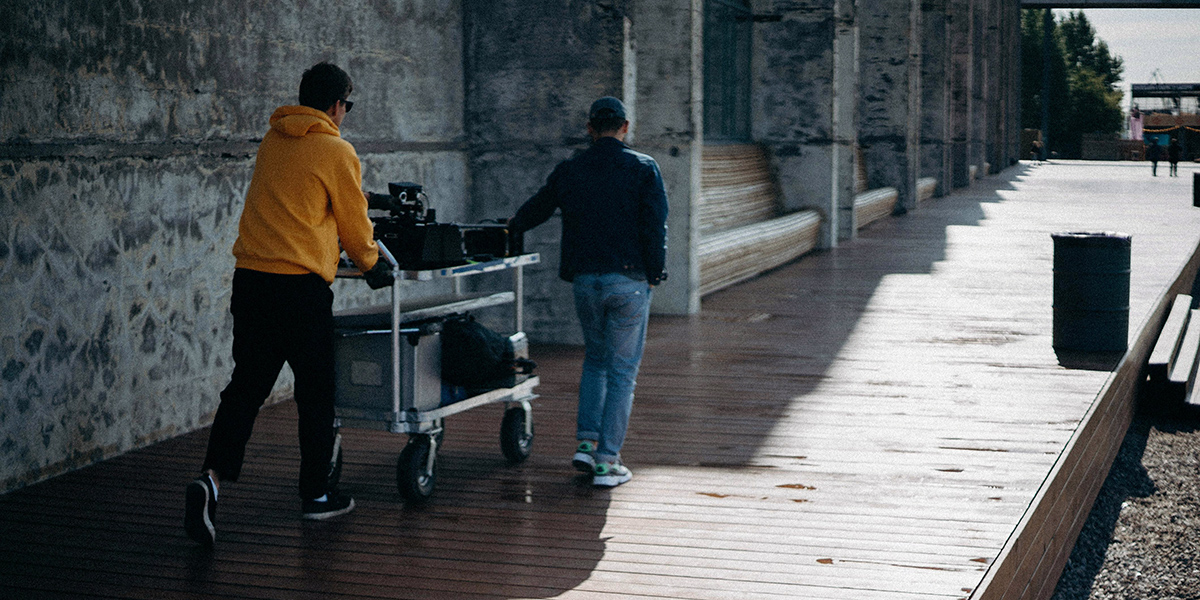 Two individuals are seen pushing a cart with equipment along a wooden path beside a concrete building in an outdoor setting.