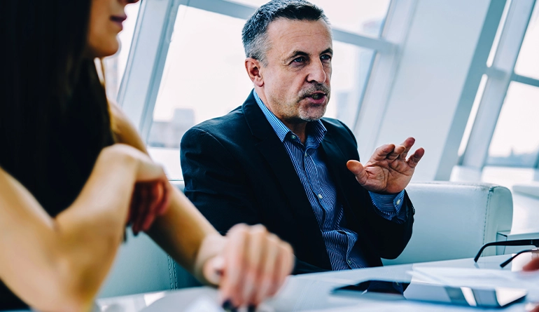 A man in a suit speaks and gestures with his hand during a business meeting at a modern office table, with another person partially visible in the foreground.