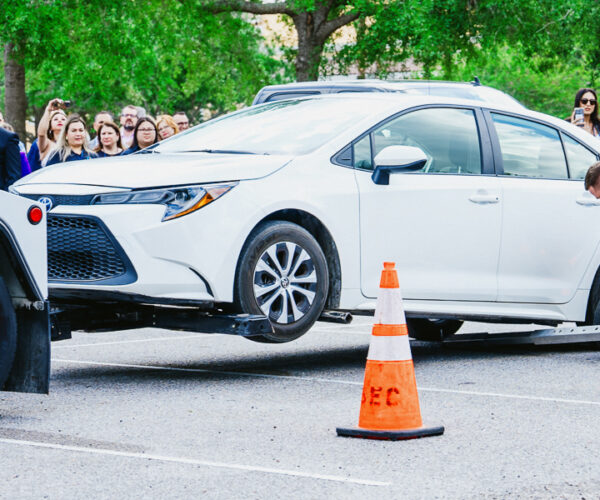 A white car is being towed in the parking lot of rosen shingle creek by a tow truck, with orange cones placed around the rear. A man is attaching the car to the truck while a crowd watches in the background.