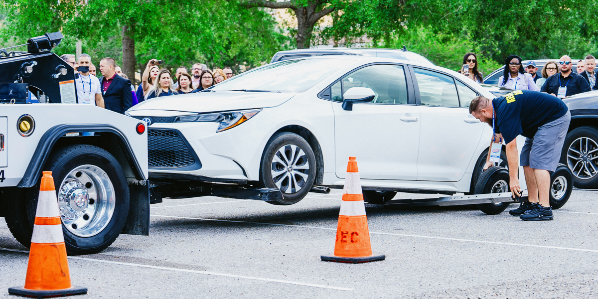 A white car is being towed in the parking lot of rosen shingle creek by a tow truck, with orange cones placed around the rear. A man is attaching the car to the truck while a crowd watches in the background.