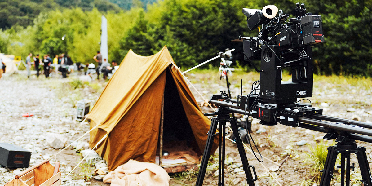 A small brown tent is set up outdoors with camera equipment on a track positioned nearby. There are people and trees in the background.