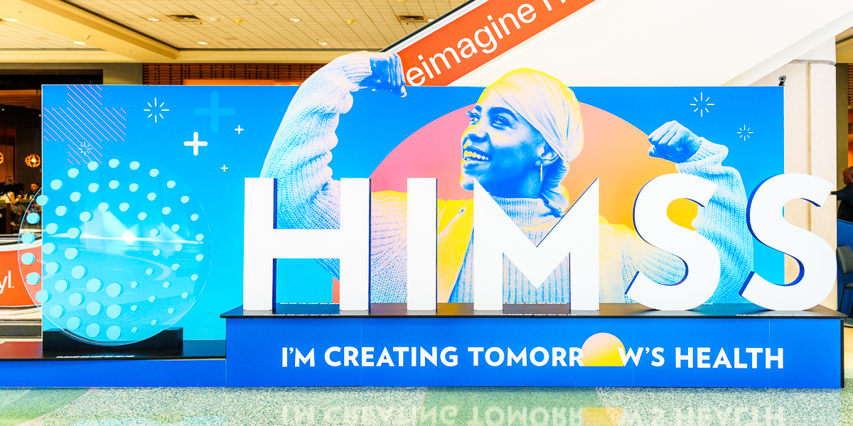 A large sign with the text "himss" and "i'm creating tomorrow's health" stands in the orange county convention center west concourse. The background displays a vibrant image of a person flexing their muscles while smiling.