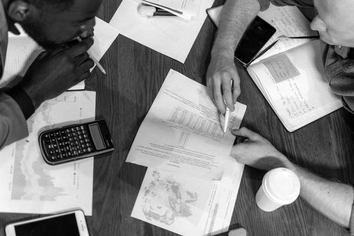 Two individuals at a wooden table discussing documents. Papers, a calculator, notebooks, a pen, a smartphone, and a coffee cup are spread out in front of them.