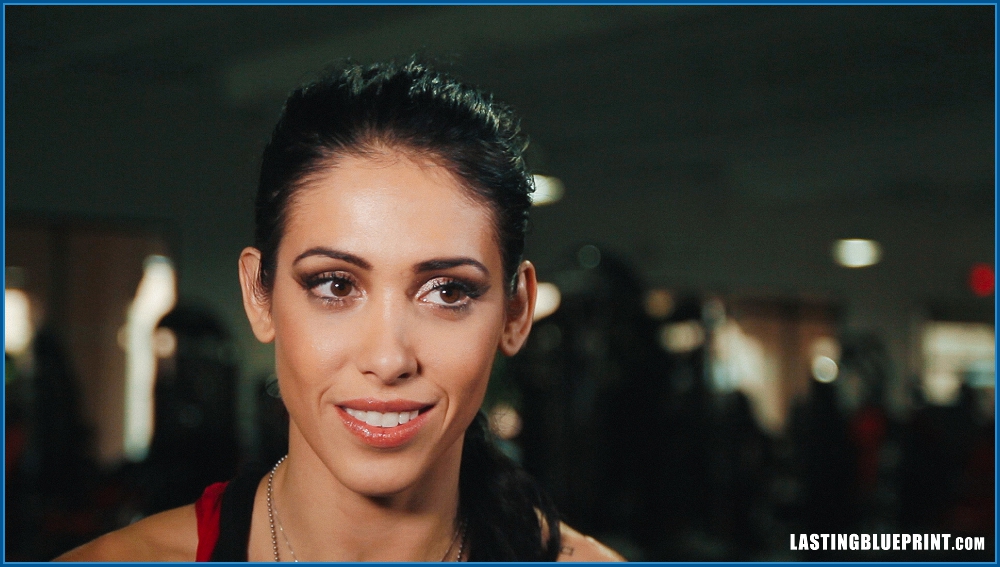 Bella falconi interview with her looking slightly to the side with a neutral expression. She is wearing a black top and a necklace, with a blurred indoor background.