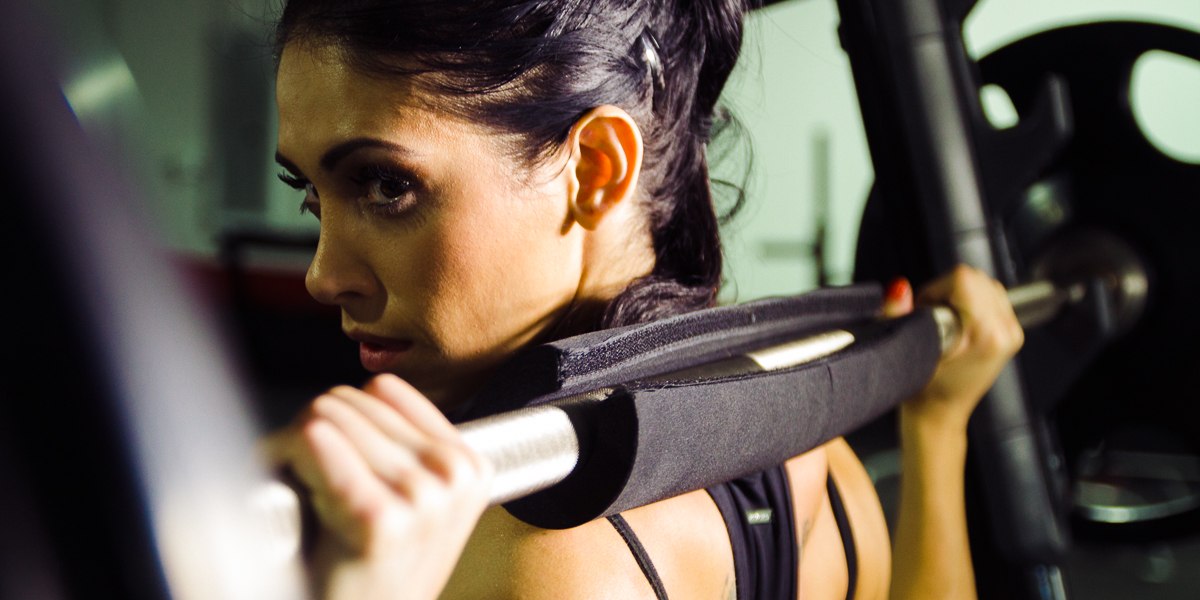 Bella falconi is holding a padded barbell on their shoulders, appearing ready to lift it. Their facial expression is focused, with a backdrop of an orlando fitness facility