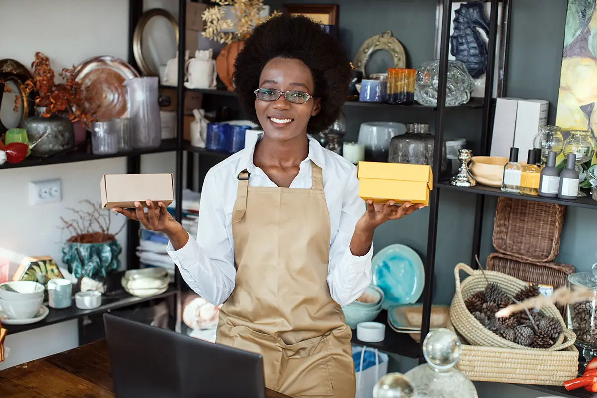 A person wearing an apron stands in front of shelves holding two boxes, one in each hand, and smiles at the camera. Various items such as plates and ornaments are displayed on the shelves behind them.