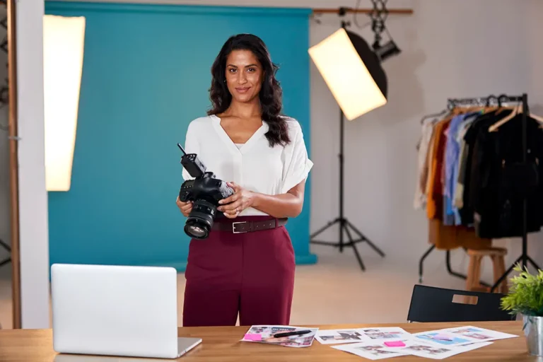 Woman holding a camera in a studio with lighting equipment, a laptop, and a clothing rack.