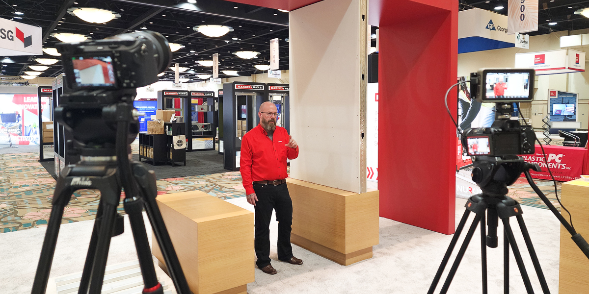 A man in a red shirt speaks in front of a camera setup at a trade show booth, filmed by an orlando videographer from lasting blueprint productions, with display panels and other booths visible in the background.