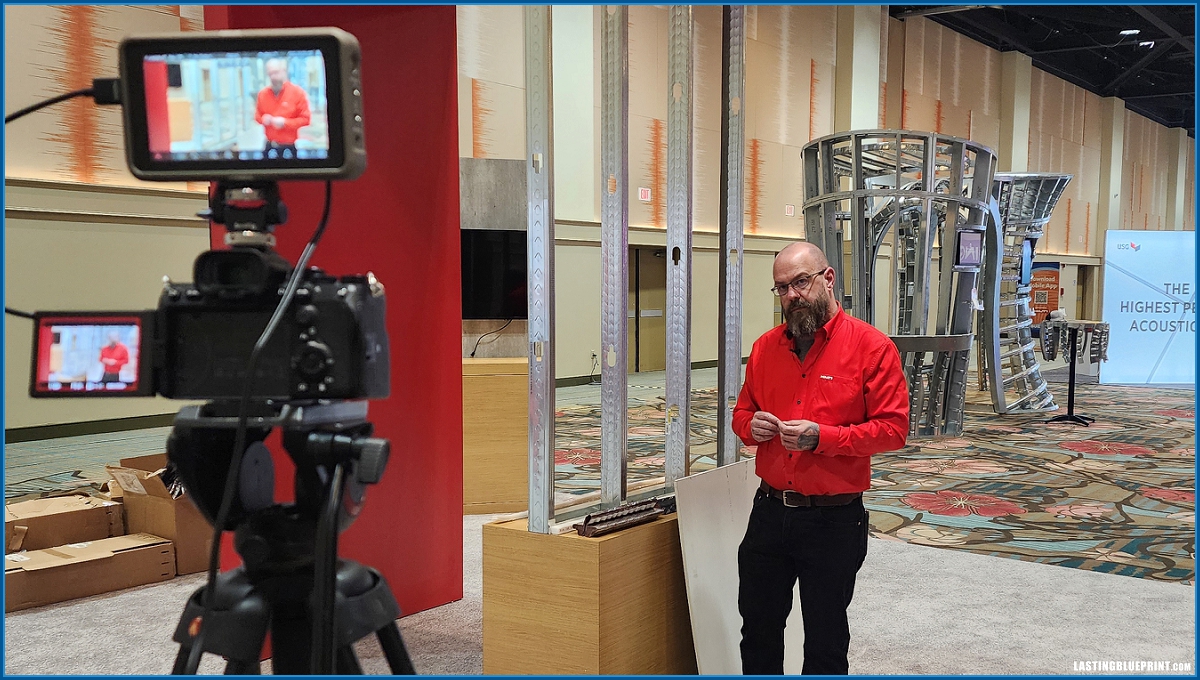 A man in a red shirt stands in front of a camera on a tripod, conducting a video recording for firestop training videos in an indoor setting with exhibit structures and posters in the background.