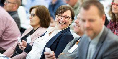 A group of people seated at a conference, some holding voting devices, with one person smiling at the camera.