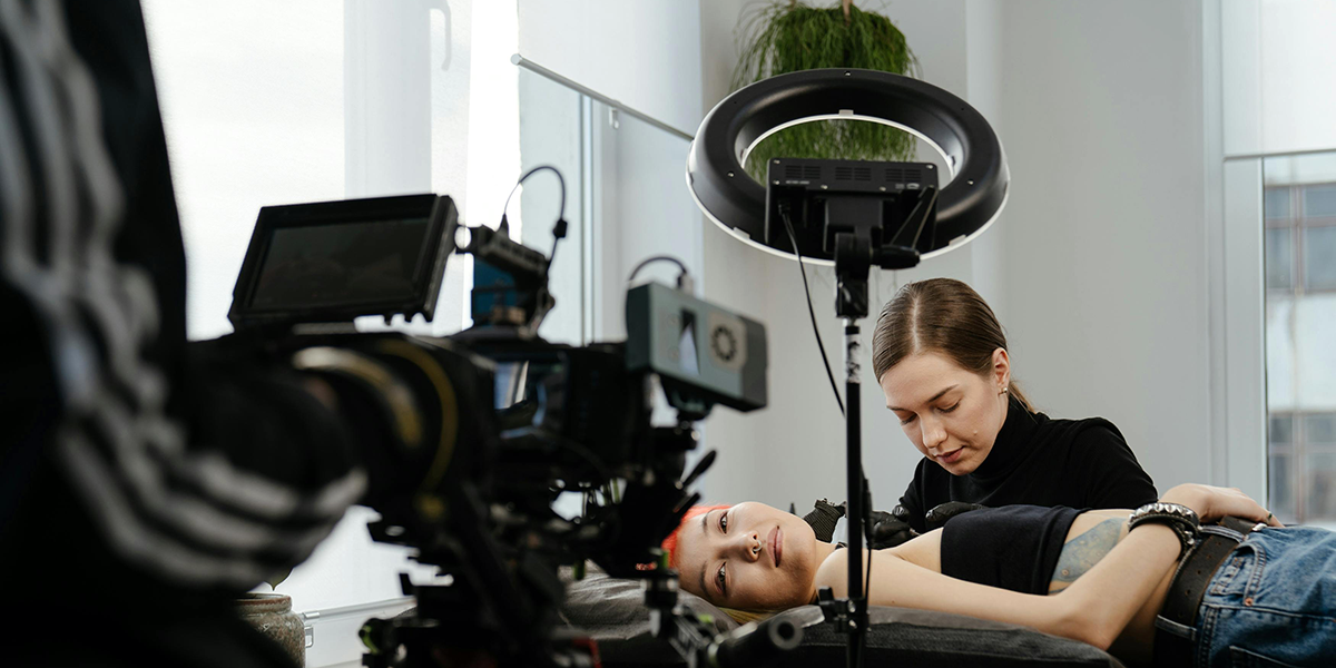 A person lies on a table while an artist, under the glow of a ring light, crafts their tattoo. Nearby, an orlando videographer captures every moment with precision.