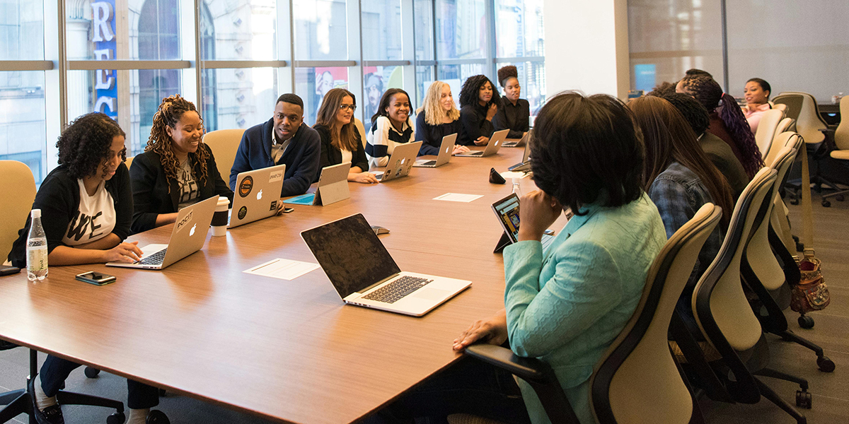 A group of people sits around a conference table with laptops in an office setting, brainstorming corporate video ideas for orlando businesses. Large windows are visible in the background, creating a bright and inspiring atmosphere for creative discussions.