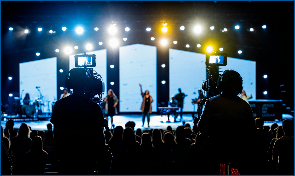 Stage performance with bright lights, silhouetted audience, and two camera operators filming the event.