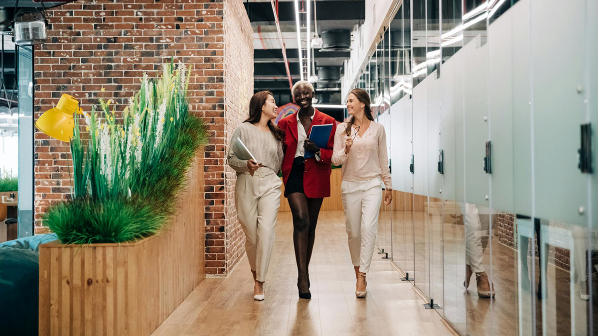 During orlando corporate video production, three people walk through a modern office hallway, chatting and smiling, embodying a vibrant, professional atmosphere. One carries a tablet and folders.