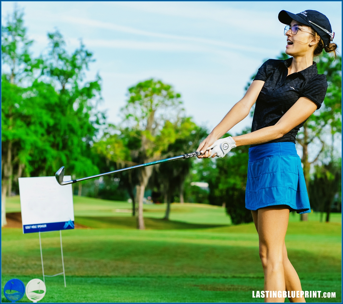 Luiza altmann, in a navy shirt and blue skirt, swings her golf club at the scenic rosen shingle creek course, surrounded by lush trees.