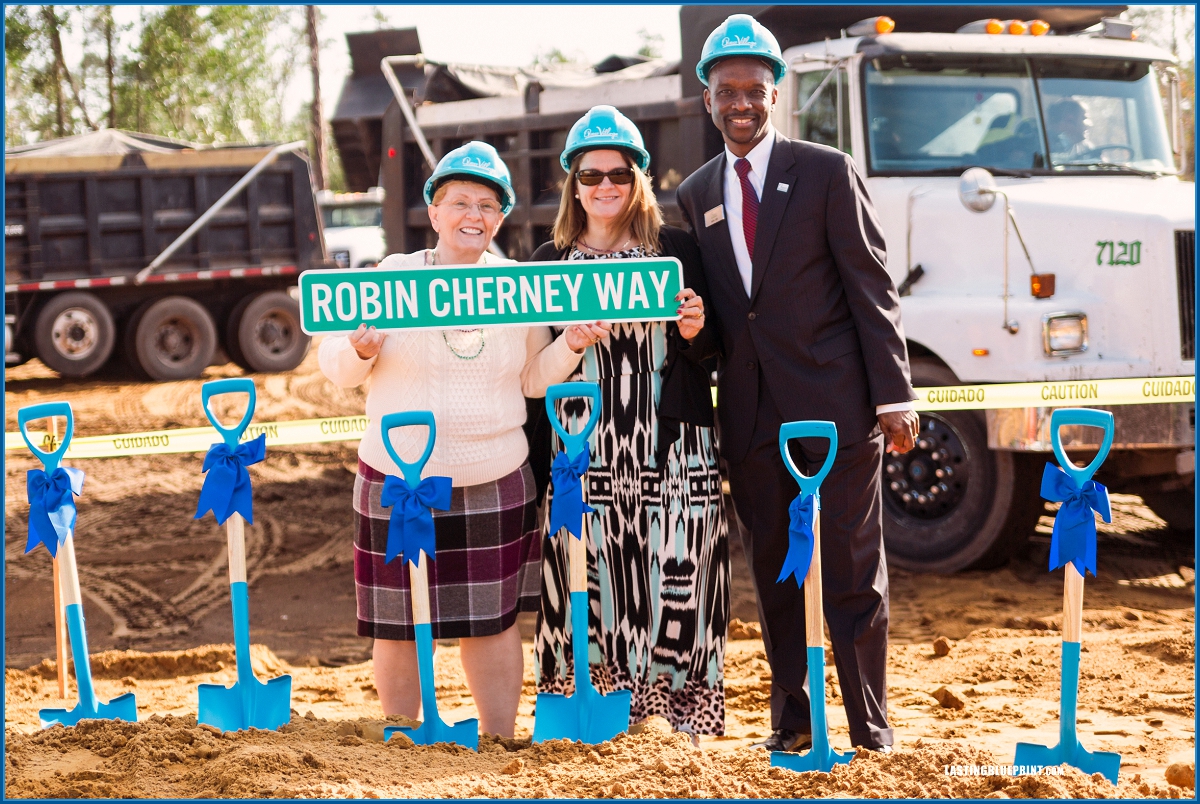 Two people holding a street sign 'robin cherney way' at quest village construction site with blue shovels, hard hats, and a truck in the background, showcasing types of event videos such as groundbreaking ceremonies.