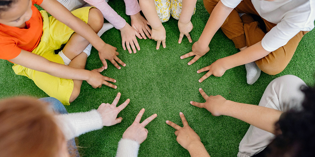 A group of children sitting on the grass, forming a circle with raised hands and peace signs.