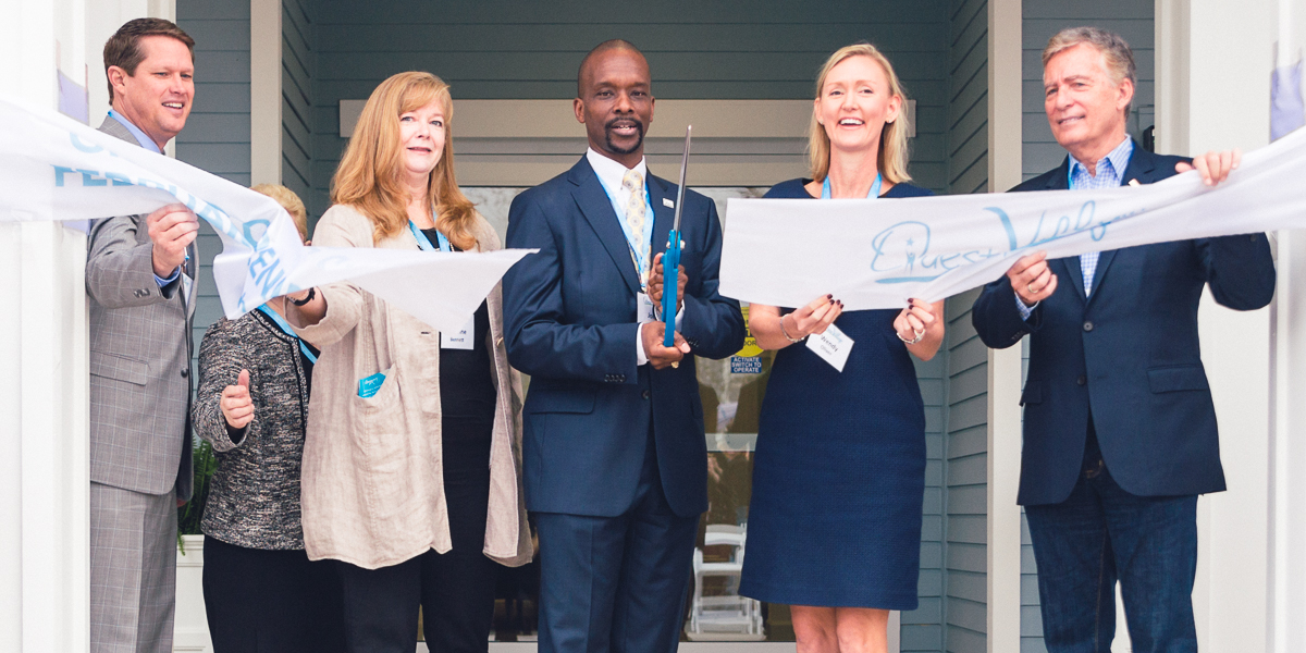 A group stands in front of quest village in orlando, cutting a ribbon with large scissors during the grand opening, while videography captures the ceremony.