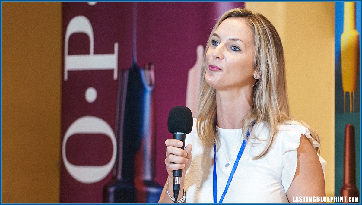 At jw marriott orlando grande lakes, a woman with a microphone speaks passionately at a beauty event, wearing a white top and blue lanyard, emphasizing the value of good testimonial questions to ask event attendees.