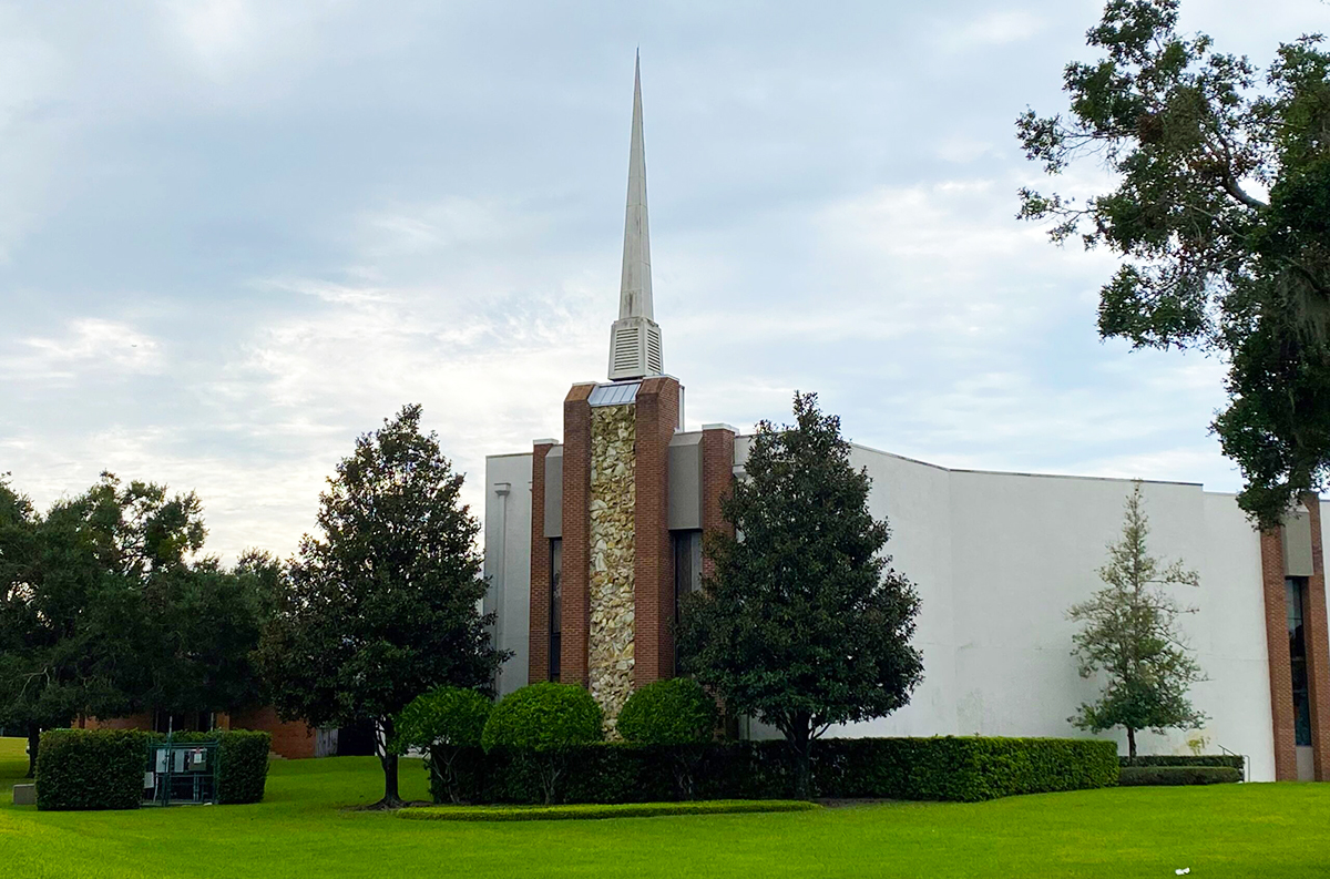 Nestled amid lush trees and grass, the trinity baptist apopka building stands proudly with its tall spire reaching into the cloudy sky.