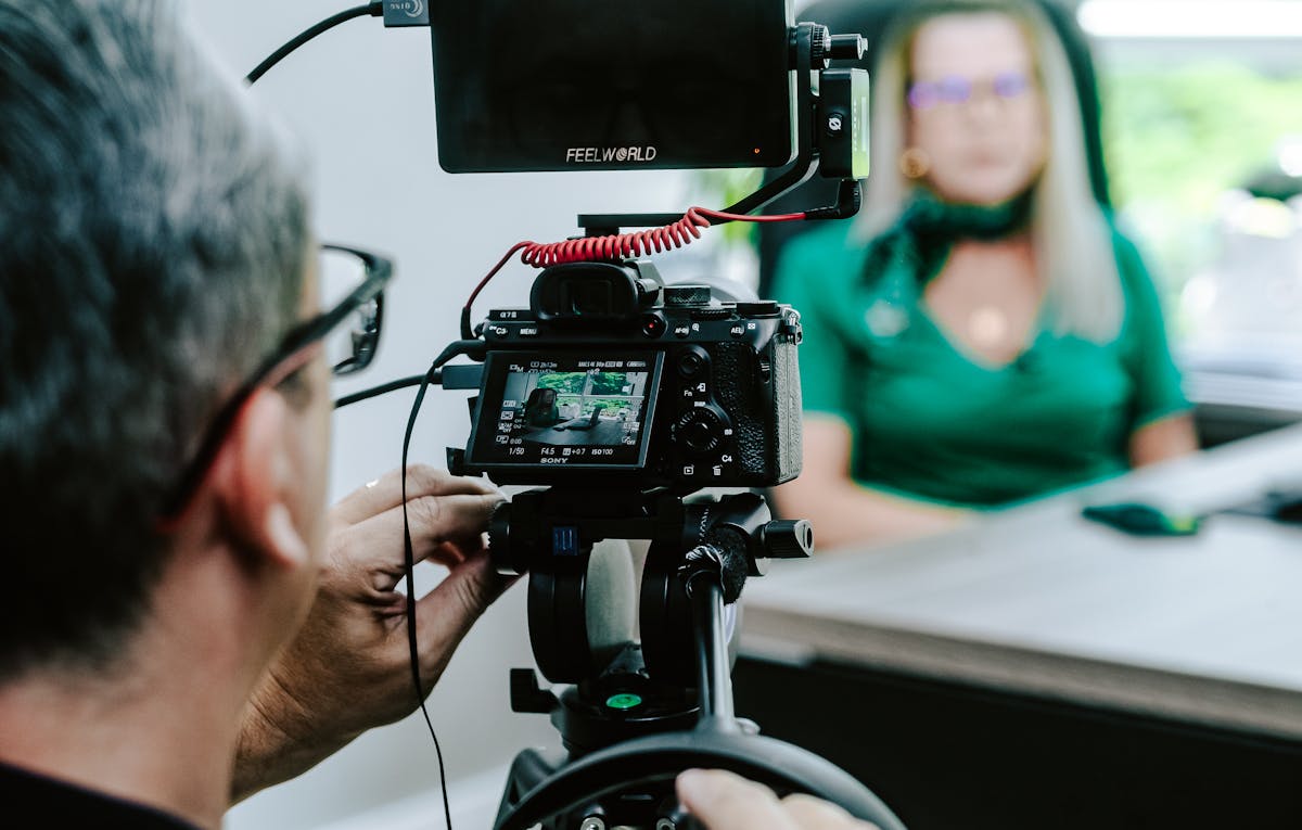 A person with short hair captures corporate videography, focusing on a woman in a green outfit seated at a table, using a professional camera setup to highlight every detail against a blurred background.