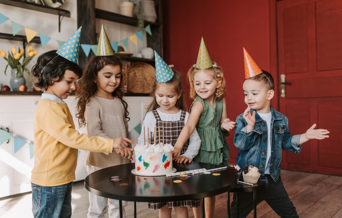 Five children wearing party hats gather around a birthday cake, with one cutting the first slice. Festive decorations brighten the scene near a red door, perfectly set for a birthday party videographer to capture every joyful moment.