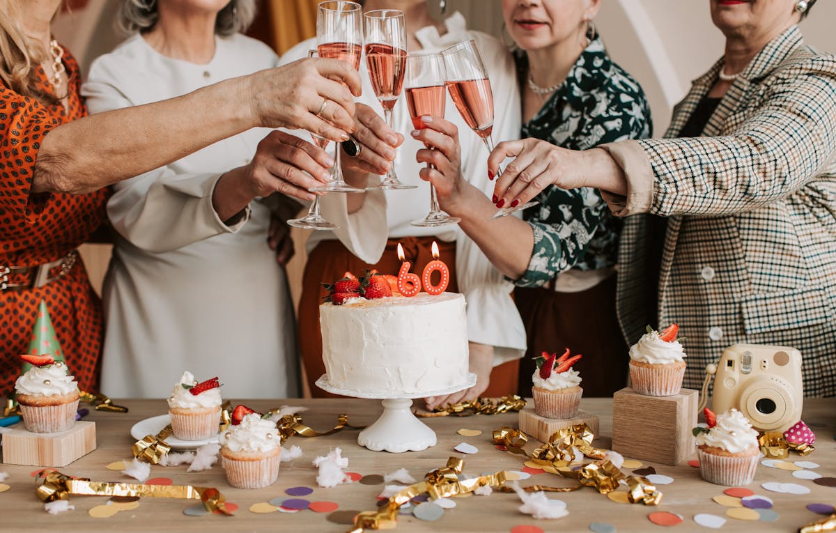 A group of people toasts with champagne glasses around a cake displaying the number 60, surrounded by cupcakes, confetti, and a birthday videographer's small camera capturing every joyful moment on the table.