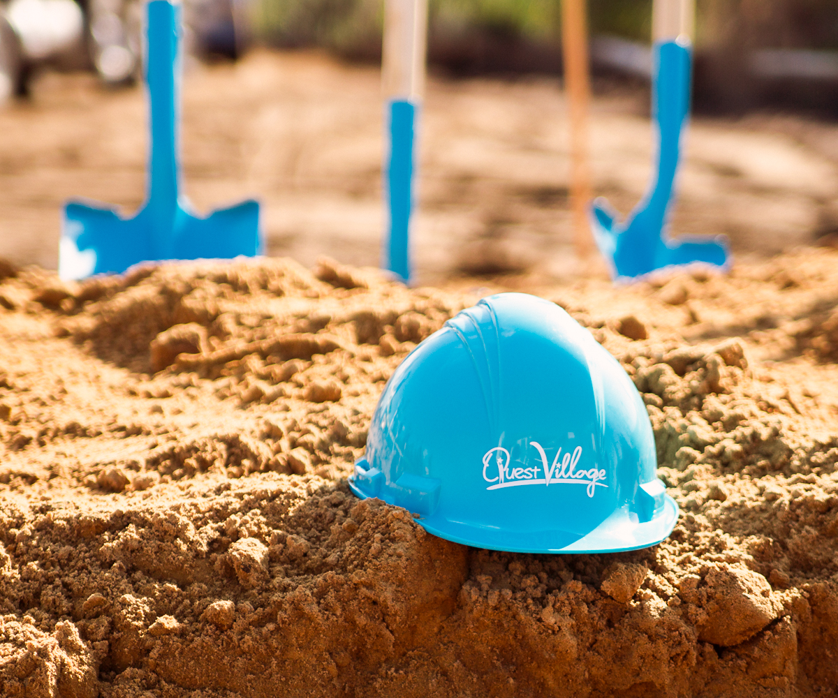 Blue hard hat on sandy ground with ceremonial shovels in the background during a groundbreaking.