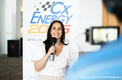 Woman speaking into a handheld microphone in front of an energy industry conference backdrop.
