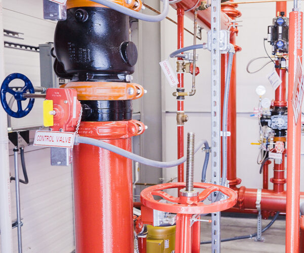 Red industrial control valve system with labeled pipes and valves, including a prominent black valve and blue handwheel, inside a facility with metal framework.