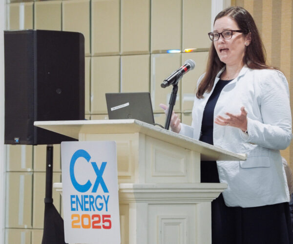 Alt text: speaker addressing the audience at a podium during the cxenergy 2025 conference, with a microphone and presentation laptop in view.