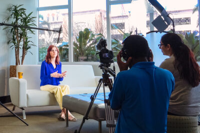 Two-camera interview setup filming a woman seated on a couch in an office.