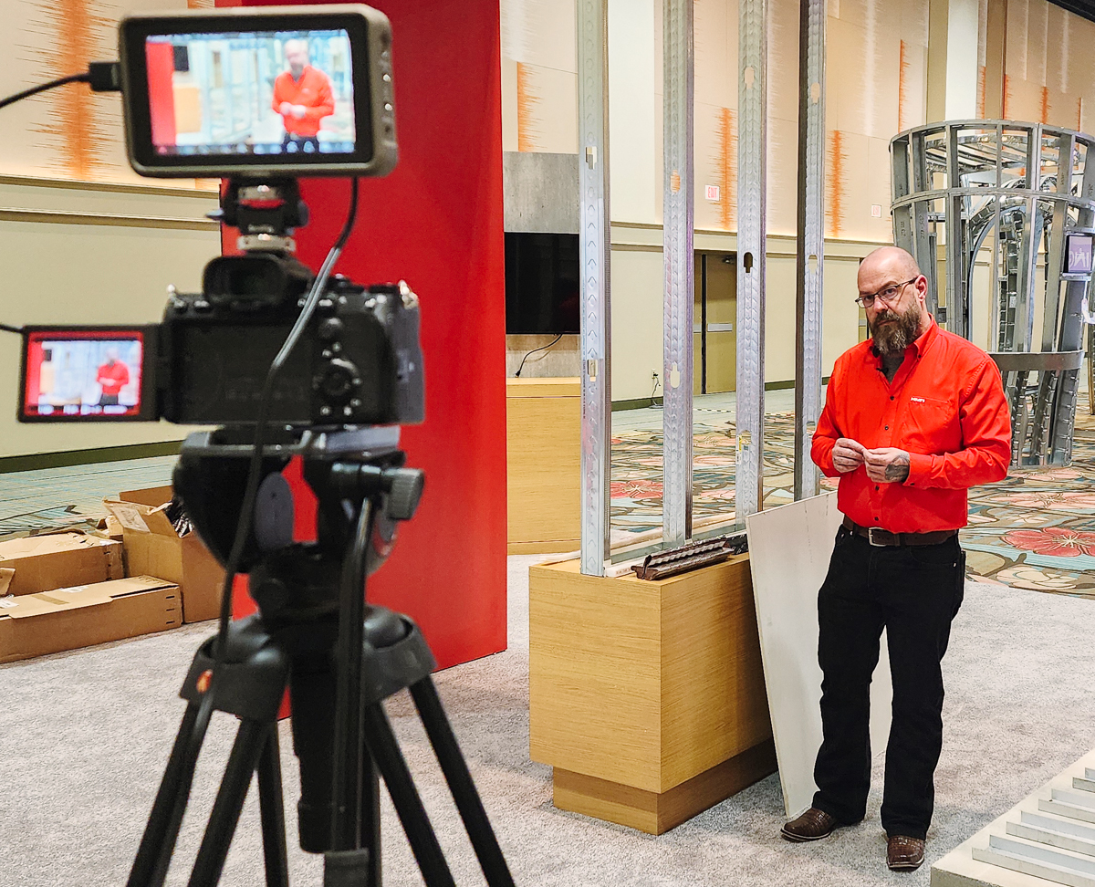 Two-camera video production setup filming a presenter beside a wall framing display.