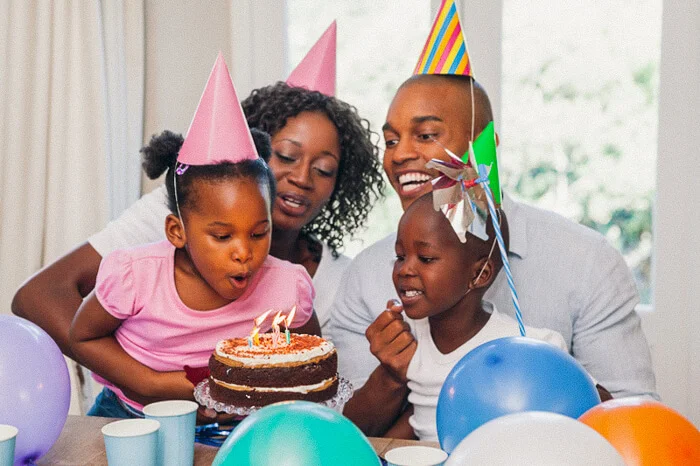 A child wearing a pink hat blows out candles on a birthday cake. Another child, an adult male, and an adult female, all wearing party hats, smile while sitting around the table adorned with balloons and cups.