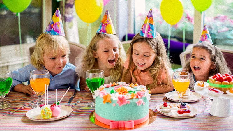 Four children wearing party hats sit around a table with a decorated cake and various desserts, celebrating a birthday.