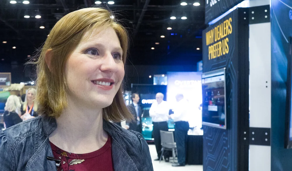 Woman smiling at a trade show, standing near a display sign reading "why dealers prefer us. " other participants and booths are visible in the background.