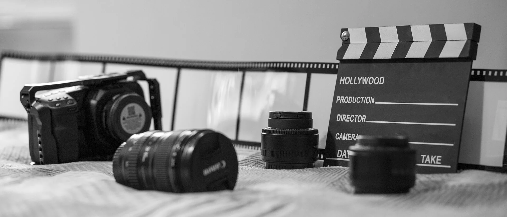 A black and white image showing a camera, several camera lenses, a filmstrip, and a clapperboard arranged on a surface.